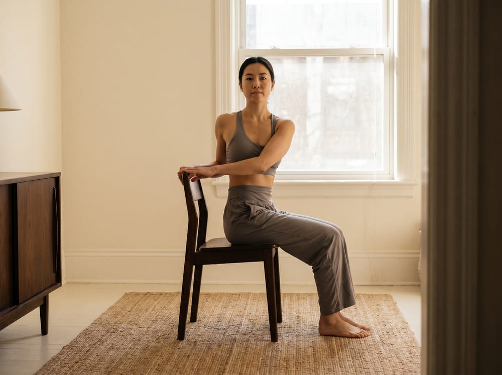 A warm, photorealistic editorial photograph in soft natural light with a warm ivory and espresso palette, 4:3 framing, no text or words anywhere: a healthy adult in their forties seated upright and fully vertical on a firm wooden chair on a mat in a bright, calm room, spine tall and the head level, performing only a small gentle turn of the upper body while the hips and head stay squarely upright and not rotated, hands resting lightly for guidance, wearing modest neutral athletic wear, soft window light across warm ivory walls.