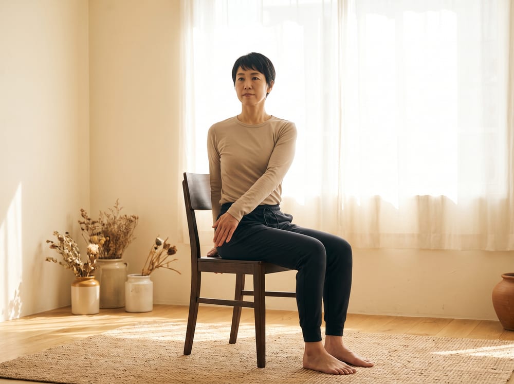 A warm, photorealistic editorial photograph in soft natural light with a warm ivory and espresso palette, 4:3 framing, no text or words anywhere: a healthy adult in their forties seated upright and fully vertical on a firm wooden chair on a mat in a bright, calm room, spine tall and the head level, performing only a small gentle turn of the upper body while the hips and head stay squarely upright and not rotated, hands resting lightly for guidance, wearing modest neutral athletic wear, soft window light across warm ivory walls.