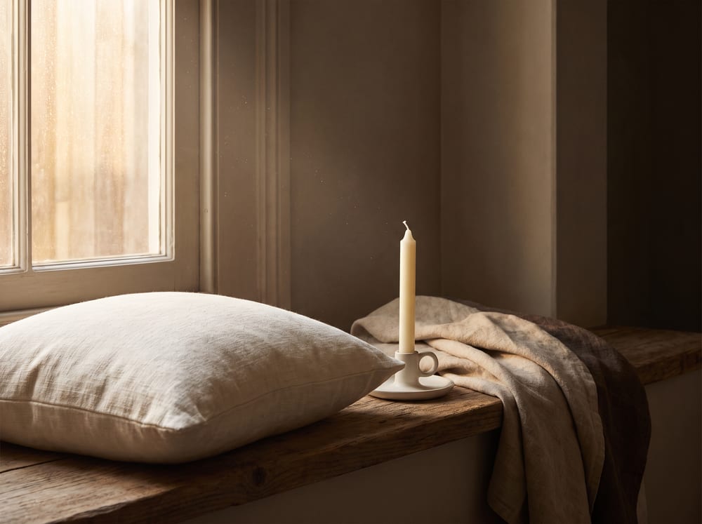 A warm, photorealistic editorial still life of a low cream-colored cushion beside a softly lit window, a single unlit ivory candle and a folded linen throw resting nearby, gentle morning light in warm ivory and espresso tones, quiet and serene, 4:3 framing, no text, no words.