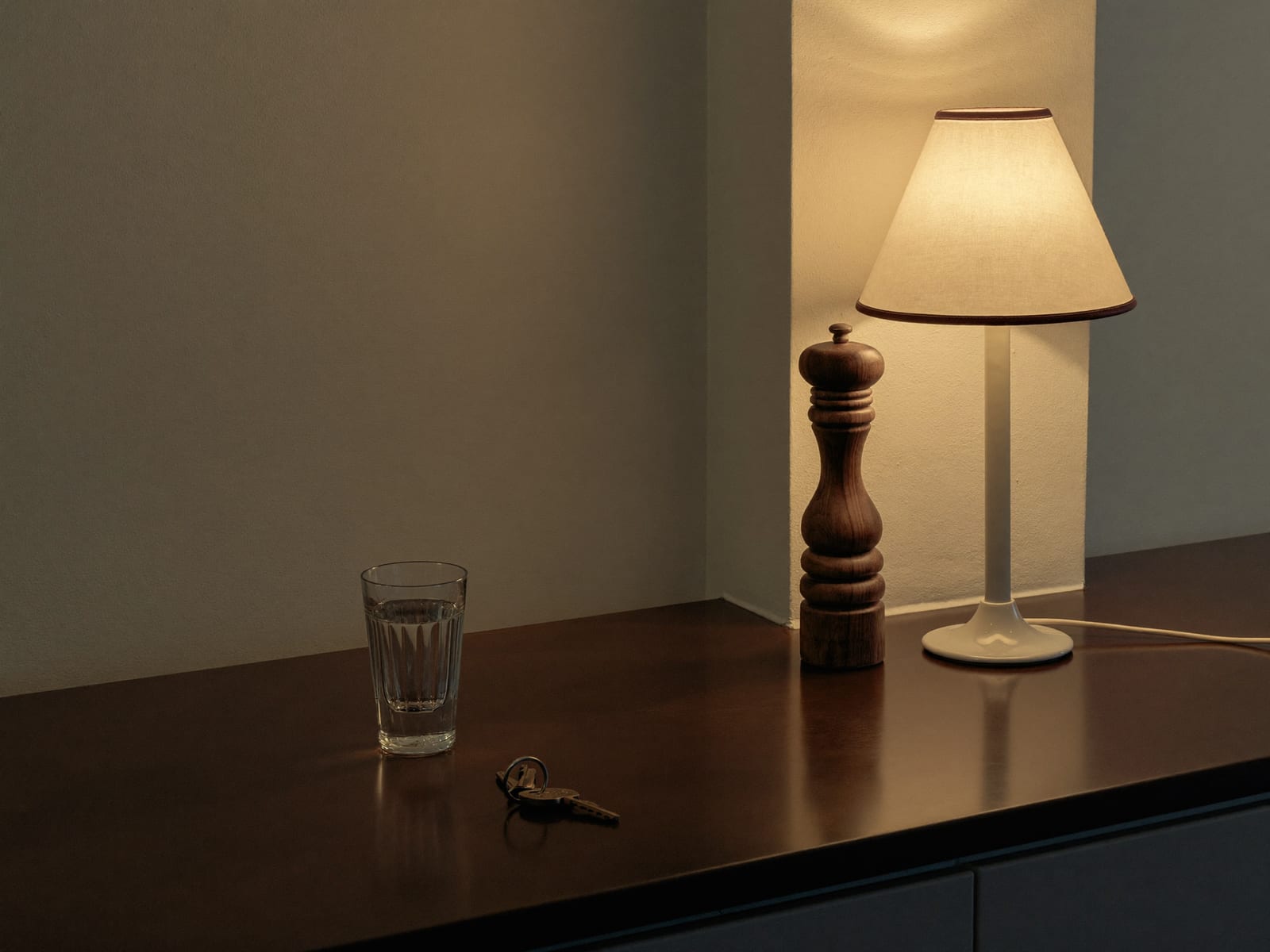 A dark wooden kitchen counter at dusk holding a glass of water, a set of house keys and a turned wooden grinder, a single lit lamp at the far end throwing warm light up an ivory plaster wall while the rest of the counter stays in shadow, nobody in the room, warm ivory and espresso palette, 4:3, no text.