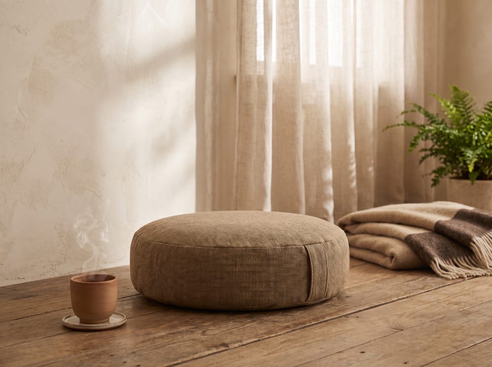 A warm, photorealistic scene in soft diffused daylight: a low meditation cushion on a wooden floor beside a small clay cup of tea, gentle window light falling across an ivory wall, warm espresso and sand tones, serene and still, 4:3 framing, no text, no words.