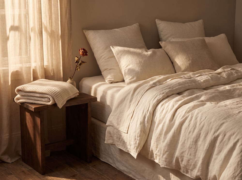 A warm, photorealistic editorial scene of a softly made bed with layered ivory linen and plush pillows, a folded cream blanket and a single dried rose stem on the nightstand, gentle late-afternoon light spilling through a sheer curtain, muted warm ivory and espresso palette, calm and restful, 4:3 framing, no text, no words.