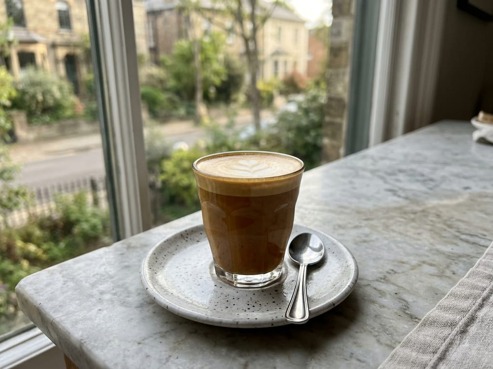 A small glass cortado with a thin cap of pale microfoam sitting over deep amber espresso, on a pale marble counter beside a few scattered coffee beans, lit by soft morning window light, shot straight on at eye level with a shallow depth of field.