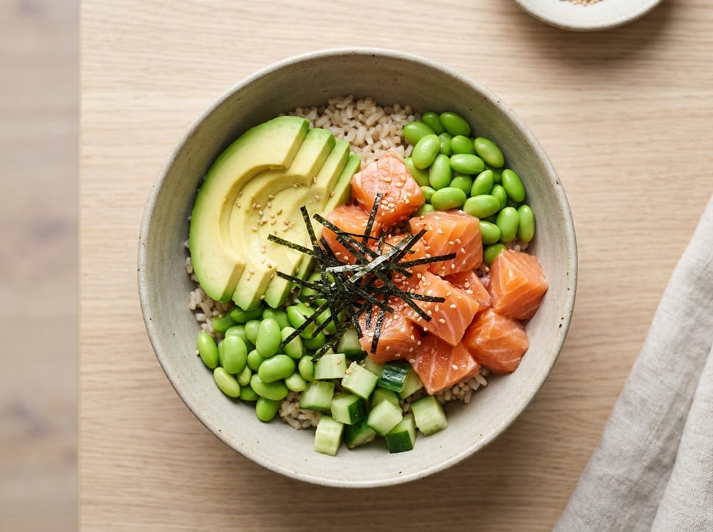 Overhead shot of a marinated raw salmon poke bowl in a matte ceramic bowl, glistening salmon cubes beside fanned avocado slices, bright green edamame, diced cucumber, thin nori strips and sesame seeds over brown rice, on a pale oak table in soft natural daylight.