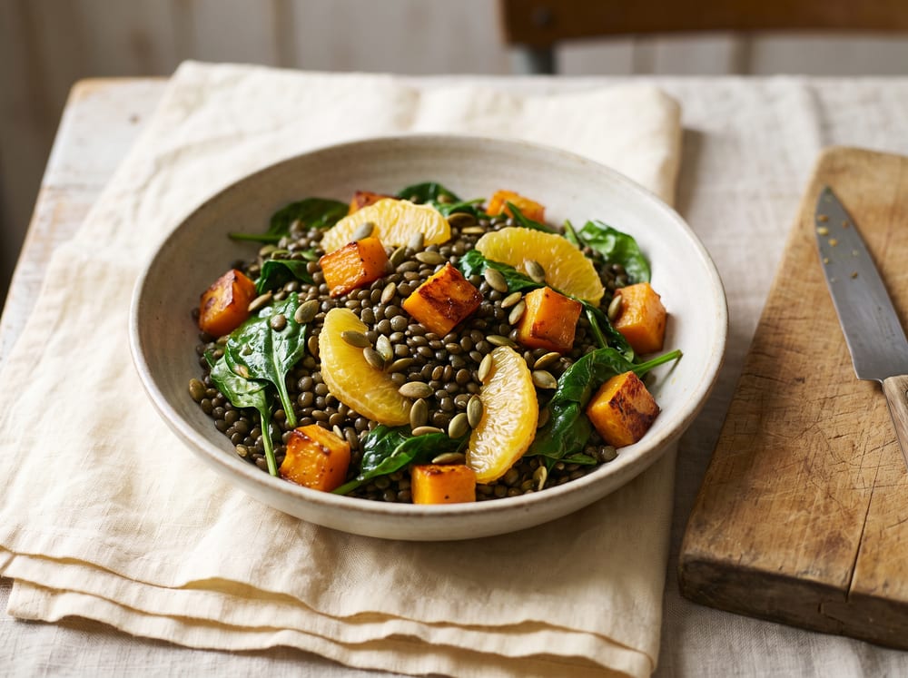 A shallow ceramic bowl of dark green lentils tossed with wilted spinach, burnished cubes of roasted butternut squash and bright orange segments, scattered with toasted green pumpkin seeds, on warm ivory linen beside a worn wooden board, soft daylight raking across from the left, photographed straight-on in a four-by-three frame with a softly blurred background, no text.