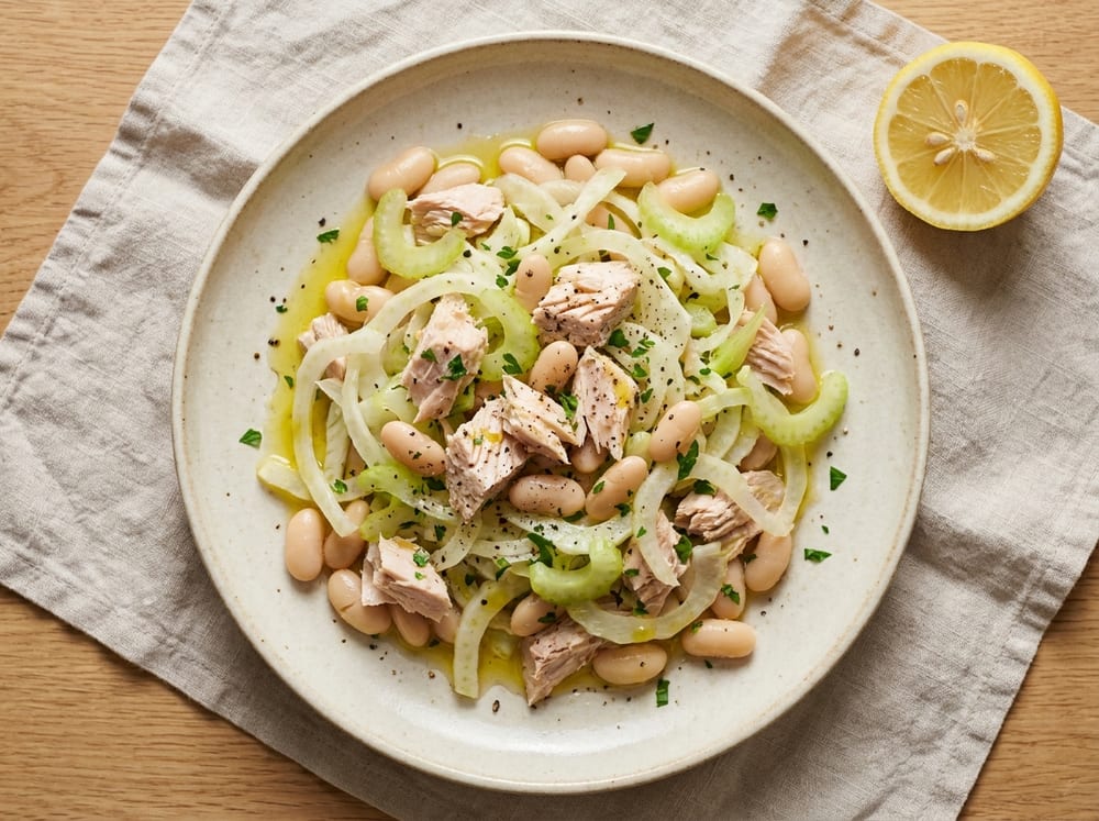 A generous plate of flaked white tuna and creamy cannellini beans tossed with pale shaved fennel and celery ribbons, resting on a bed of dark green arugula, glistening with olive oil and speckled with parsley and black pepper, a cut lemon half beside it, shot top-down on a cream stoneware plate atop a light linen napkin in bright natural window light.