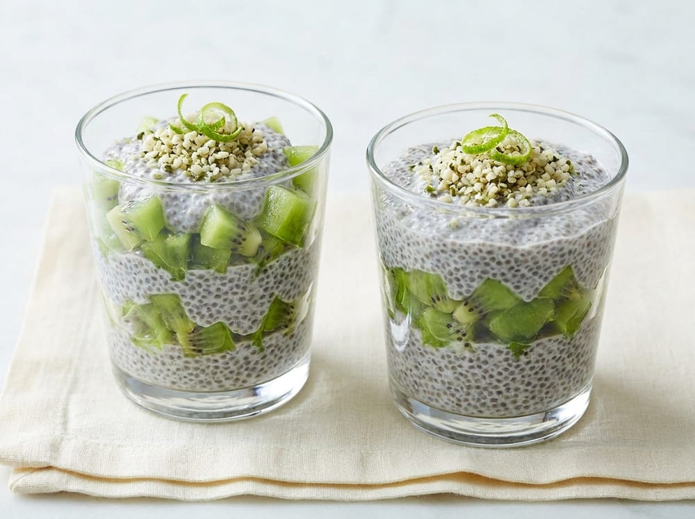 Two small clear glasses on pale ivory linen holding layers of speckled grey vanilla chia pudding and bright emerald diced kiwi, crowned with a scatter of soft cream-and-green hemp hearts and a wisp of lime zest, cool and glossy under soft diffused daylight, shot slightly above eye level.
