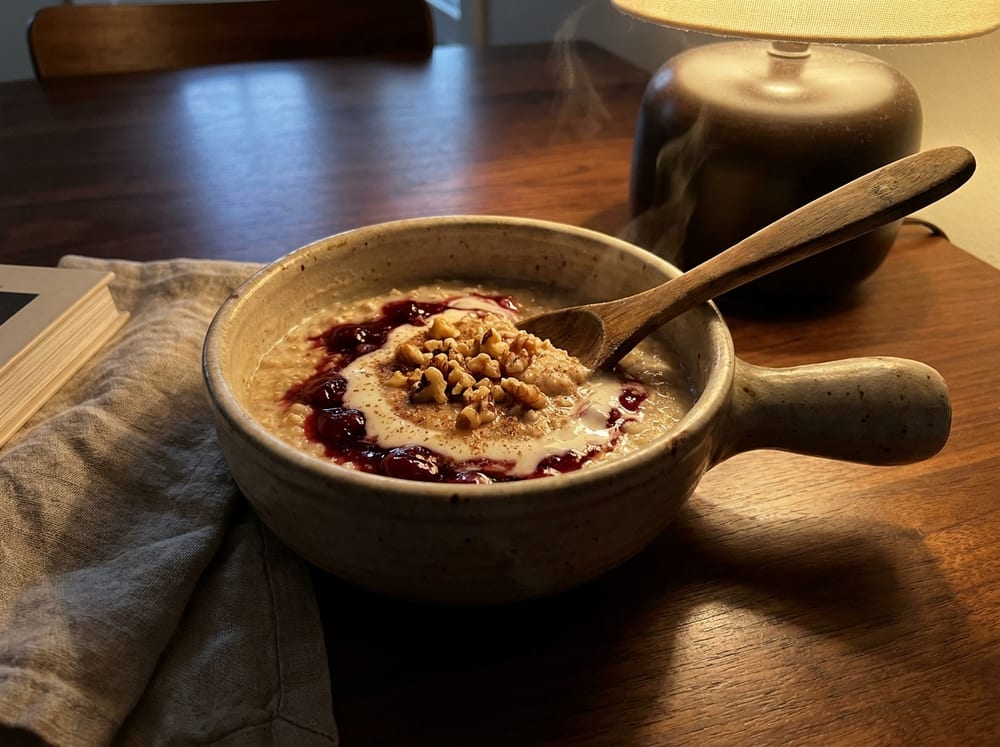 A small ceramic spoon-pot of warm oats swirled deep ruby with tart cherry compote, a pale ribbon of kefir marbled through the surface, chopped walnuts and a dusting of nutmeg on top, resting on a dark walnut table under soft warm low-angle evening lamplight with a linen napkin just out of focus